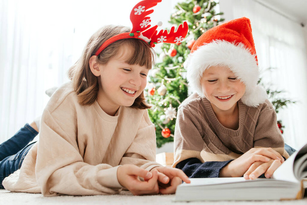 Happy children, a boy and a girl, read a book on Christmas or New Years Eve against the background of a Christmas tree. Waiting for the holidays. - Photo, Image
