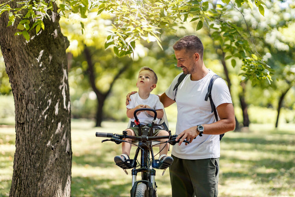 A puzzled son looking at the tree and sitting on a bike while father explaining him photosynthesis. - Photo, Image