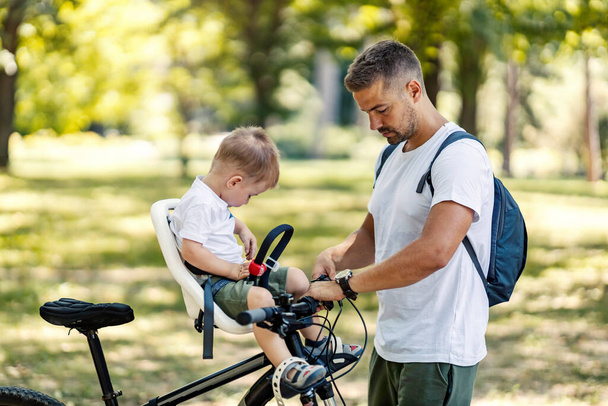 Father buckling his son's seat and preparing to healthy bike ride in forest. - Photo, Image
