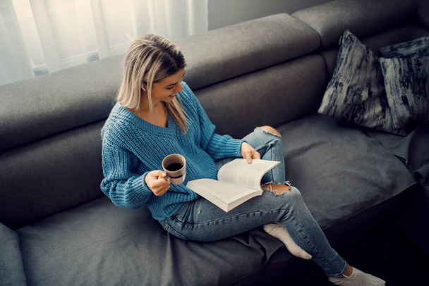Szczęśliwa kobieta pije kawę i czyta książkę, relaksując się w przytulnym domu. - Zdjęcie, obraz