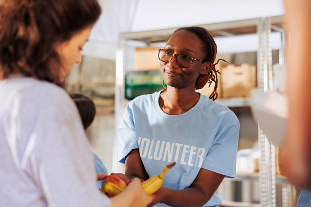 Imagen detallada que muestra a la voluntaria afroamericana ofreciendo comidas gratis a los pobres y necesitados. Niza mujer negra en un banco de alimentos al aire libre compartiendo productos frescos y alimentos para los desfavorecidos. - Foto, imagen