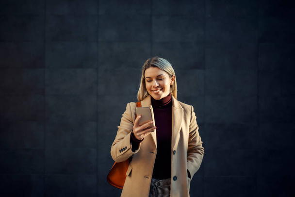 Una mujer con abrigo parada al aire libre y sonriendo al teléfono. - Foto, Imagen