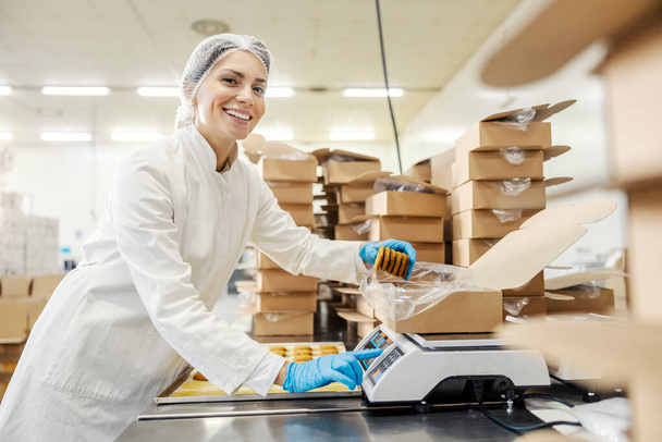 Un trabajador de una planta de alimentos está midiendo las galletas y poniéndolas en una caja mientras sonríe a la cámara. - Foto, imagen