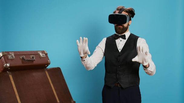 Moderner Portier mit Vr-Brille vor blauem Hintergrund, der Virtual-Reality-Technologie am Headset im Studio nutzt. Junger Kellner mit edlem Beruf steht neben Einkaufstaschen, Reisebranche. - Foto, Bild
