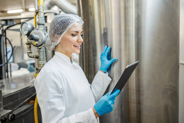 Ein Arbeiter einer milchverarbeitenden Fabrik kontrolliert eine Maschine mit Milch, während er Tabletten in der Hand hält und sie anlächelt. - Foto, Bild
