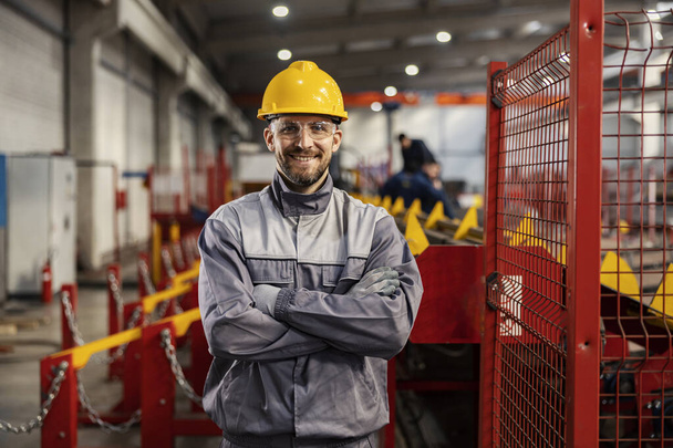 Dumny metalurg i pracownik przemysłu ciężkiego stoi w fabryce z skrzyżowanymi ramionami i uśmiecha się do kamery. - Zdjęcie, obraz