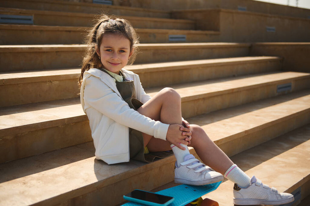 Caucasian elementary age child, cute little kid girl with skateboard and smart mobile phone, sitting on steps outdoors and smiling looking at camera. People. Active lifestyle. Happy carefree childhood - Photo, Image