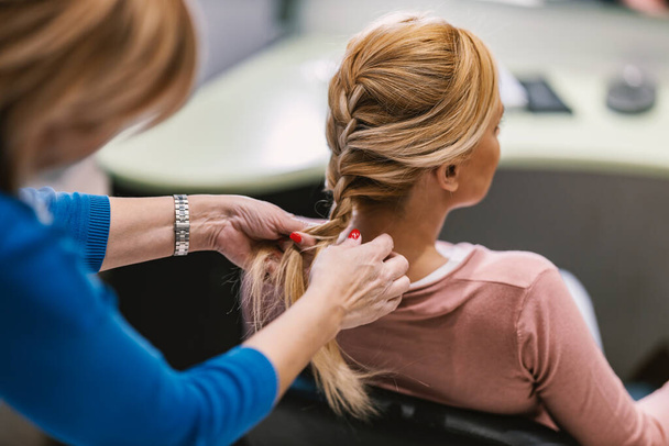 Vue arrière d'une femme blonde assise dans un salon pendant qu'un coiffeur fait une tresse. - Photo, image