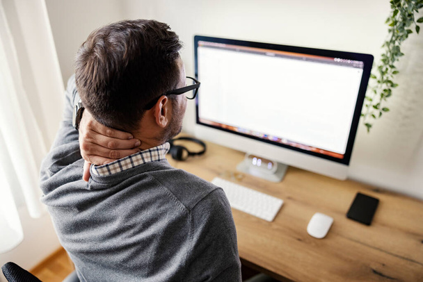 Πίσω όψη ενός εργαζομένου κάθεται στο γραφείο στο σπίτι και τρίβει τον πονεμένο λαιμό του. - Φωτογραφία, εικόνα