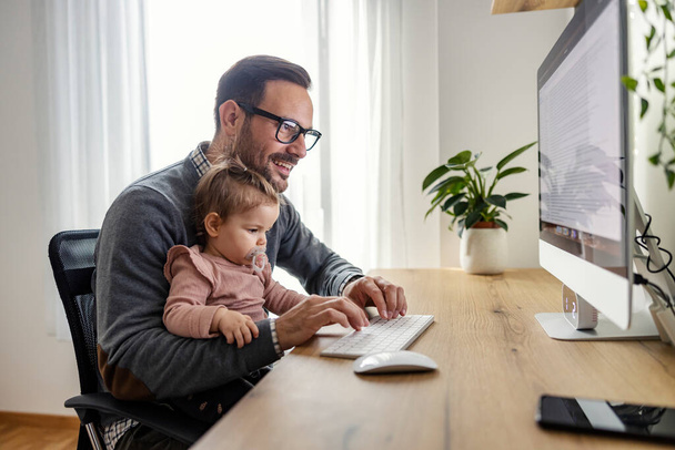 Un padre impegnato sta tenendo la sua bambina in grembo mentre finisce il rapporto su un computer in ufficio. - Foto, immagini