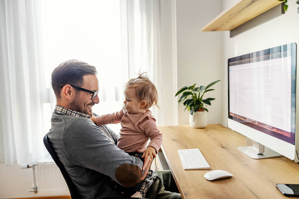 Un empresario está jugando con su bebé mientras está sentado en la oficina en casa en un descanso de trabajo. - Foto, imagen
