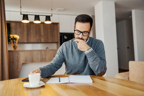 Ένας εστιασμένος επιχειρηματίας σκέφτεται και κοιτάζοντας το χρονοδιάγραμμα, ενώ πίνοντας καφέ στο σπίτι. - Φωτογραφία, εικόνα
