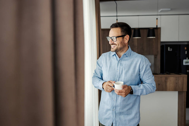 Un homme souriant regarde par la fenêtre tout en tenant du café frais du matin. - Photo, image