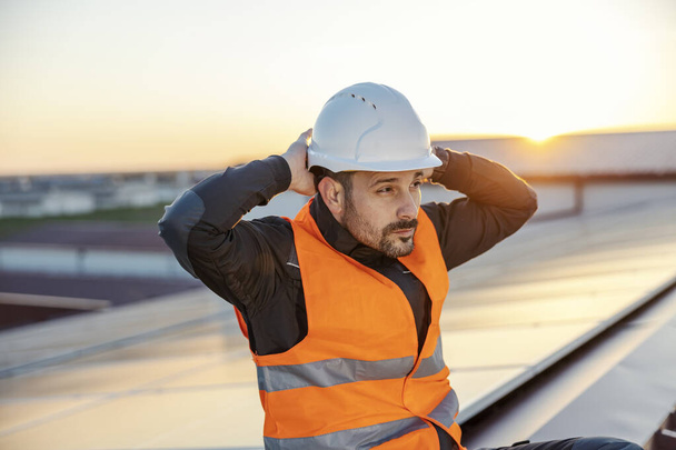 Werknemer zet helm op zijn hoofd en bereidt zich voor op het werk terwijl hij op het dak naast de zonnepanelen zit. - Foto, afbeelding