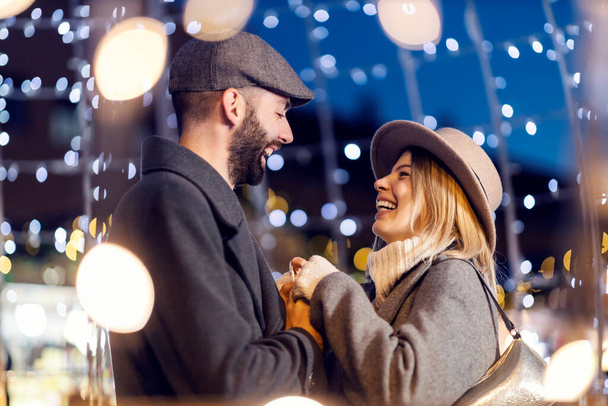 Karácsonyi pár fogja egymás kezét a szabadban, karácsonyi fényekkel körülvéve szilveszterkor. Egy fiatal, szerelmes pár áll az utcán, fogják egymás kezét, és szilveszterkor szemkontaktust létesítenek.. - Fotó, kép