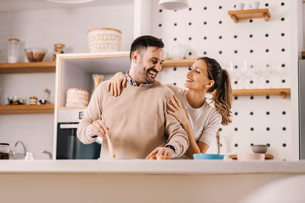 Ein glücklicher Mann frühstückt zu Hause in der Küche, während eine Frau ihn umarmt. - Foto, Bild