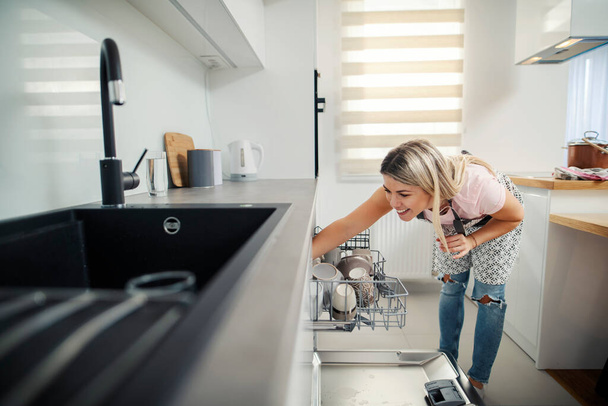 Eine Frau in der heimischen Küche legt Geschirr in die Geschirrspülmaschine. - Foto, Bild