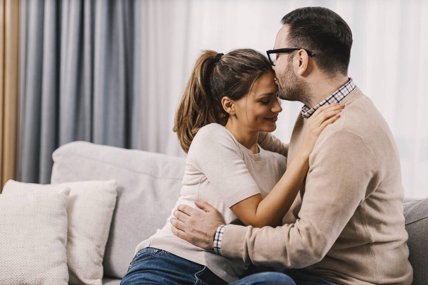 Egy férj, aki élő kapcsolatot mutat a feleségével, és homlokon csókolja, miközben otthon ül.. - Fotó, kép