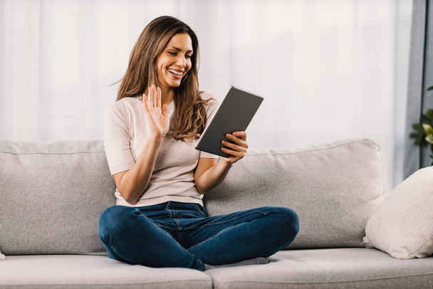 Una donna si siede in salotto, ha una videochiamata, sorride e saluta il tablet.. - Foto, immagini