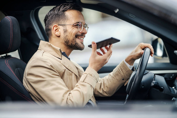 Rušný mladý inteligentní ležérní muž řídí své auto a používá svůj telefon pro hlasovou zprávu. - Fotografie, Obrázek