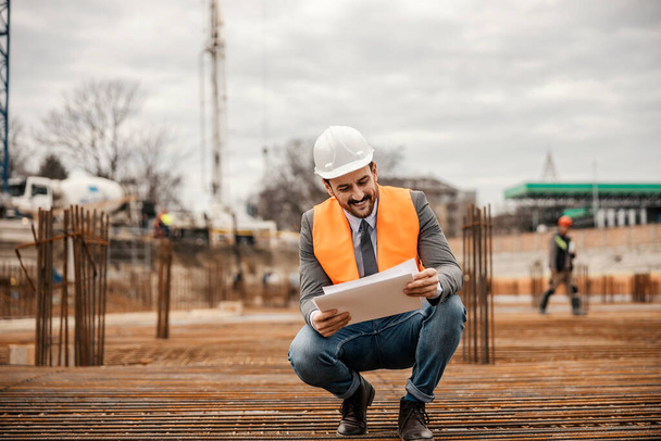 Un hombre de negocios alegre se agacha en el área de reconstrucción en la construcción de cimientos y mirando documentos y planos. - Foto, Imagen
