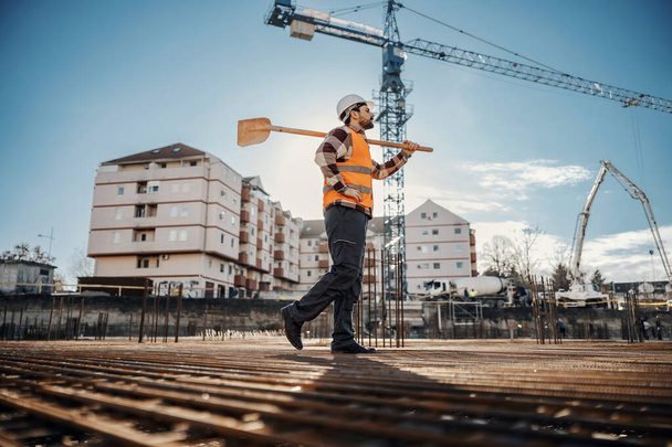 Трудолюбивый строитель с лопатой на плече ходит по площадке на фундаменте здания и готовится к работам. - Фото, изображение