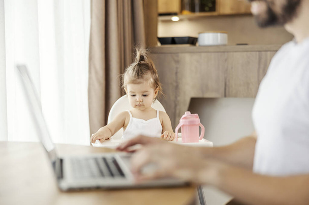 Portret van een schattig klein meisje in haar stoel, terwijl vader is met behulp van laptop voor werk en typen op het toetsenbord. - Foto, afbeelding