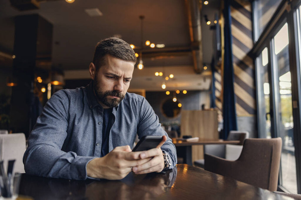 Ένας ενοχλημένος άντρας κάθεται στο καφέ και διαβάζει άσχημα νέα στο τηλέφωνο.. - Φωτογραφία, εικόνα