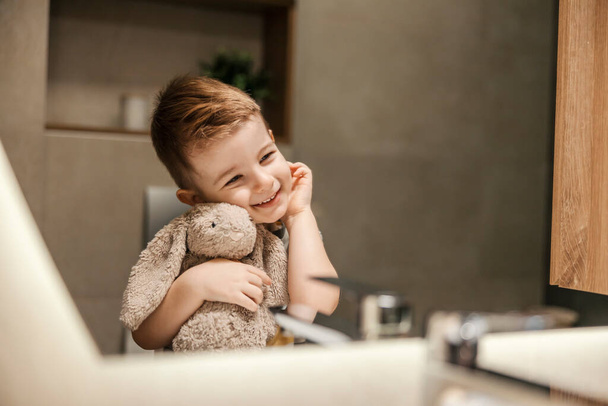 An adorable little boy is smiling at his reflection in the mirror in the bathroom and hugging a bunny after morning routine. - Photo, Image