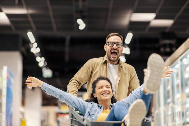 Ένας ευτυχισμένος άντρας οδηγεί τη γυναίκα του σε ένα καροτσάκι στο σούπερ μάρκετ και διασκεδάζει.. - Φωτογραφία, εικόνα