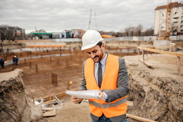 Veselý šéf staveniště stojí na staveništi a prohlíží si plány a dokumenty. - Fotografie, Obrázek