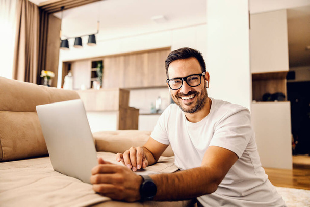 Een man zit thuis internet te zoeken op laptop en lacht naar de camera. - Foto, afbeelding