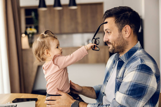 Дочь играет с отцовскими очками, сидя дома на столе в рабочее время. - Фото, изображение