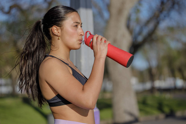 Portret van latijns meisje in sportkleding drinkwater uit een rode fles. - Foto, afbeelding