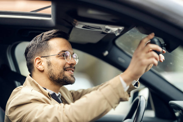 Um empresário feliz no casual inteligente está ajustando espelho retrovisor em um carro. - Foto, Imagem