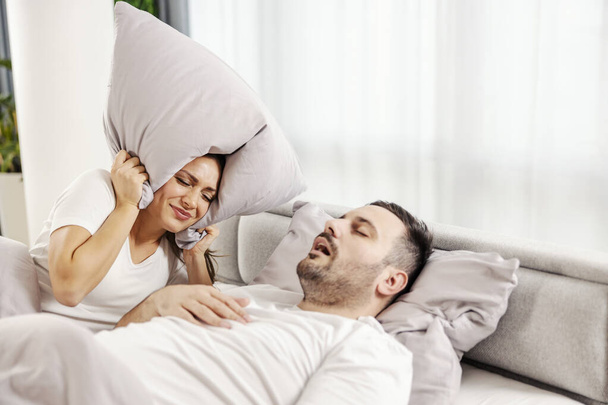 Wkurzona kobieta zakrywa uszy poduszką, podczas gdy jej mąż śpi głęboko i chrapie.. - Zdjęcie, obraz