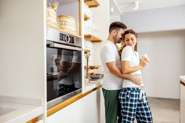 Ein liebevolles Paar trinkt zu Hause in der Küche Kaffee, während es kuschelt und sich umarmt. - Foto, Bild