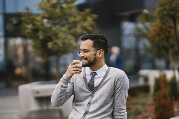 Hymyilevä nuori liikemies istuu ulkona ja siemailee kahvia kahvitauolla.. - Valokuva, kuva