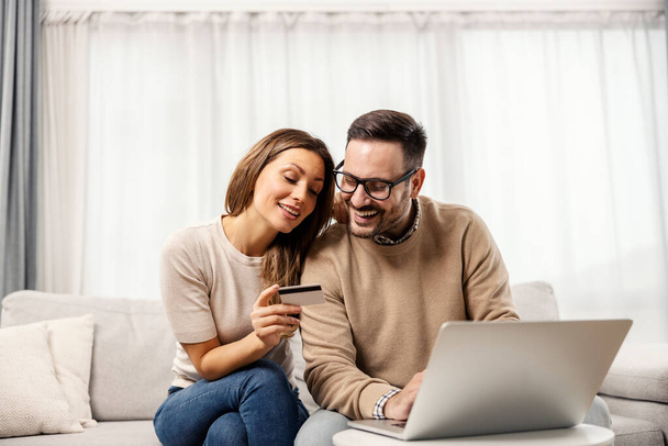 Το στοργικό ζευγάρι κάθεται σε ένα σαλόνι στο σπίτι και αγοράζει πράγματα σε απευθείας σύνδεση σε ένα φορητό υπολογιστή. - Φωτογραφία, εικόνα