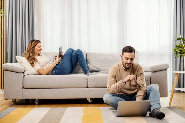 Ein glückliches Paar nutzt Tablet und Laptop zu Hause im Wohnzimmer. - Foto, Bild