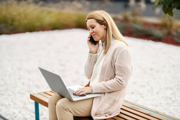 Una mujer de negocios en casual inteligente está sentada en un banco en el parque, teniendo una conversación telefónica en el teléfono y escribiendo informe en un ordenador portátil. - Foto, imagen
