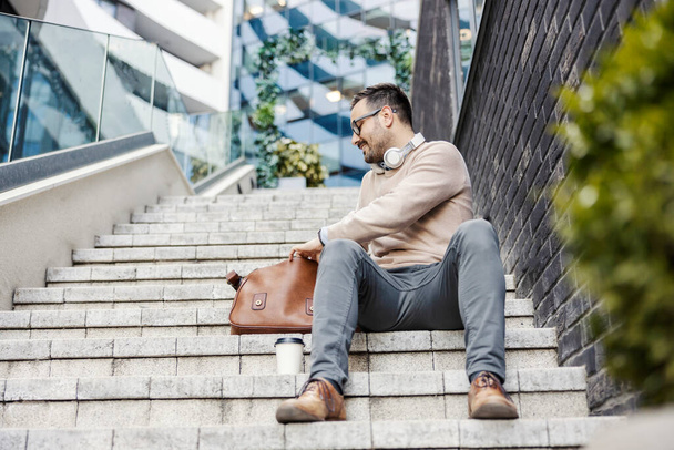 Un hombre de negocios casual inteligente se sienta en las escaleras frente al centro de negocios y busca documentos en su bolso. - Foto, imagen