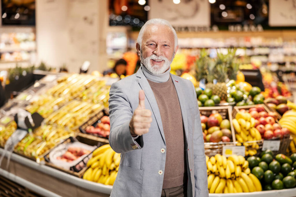 Um homem idoso feliz está dando os polegares para cima enquanto olha para a câmera no supermercado. - Foto, Imagem