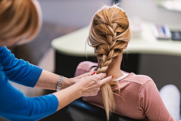 Back view of a client sitting in a salon while hairdresser is making braid. - Photo, Image
