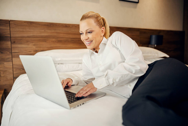 Szczęśliwa bizneswoman w podróży służbowej pracuje nad projektem leżąc na łóżku w pokoju hotelowym. - Zdjęcie, obraz