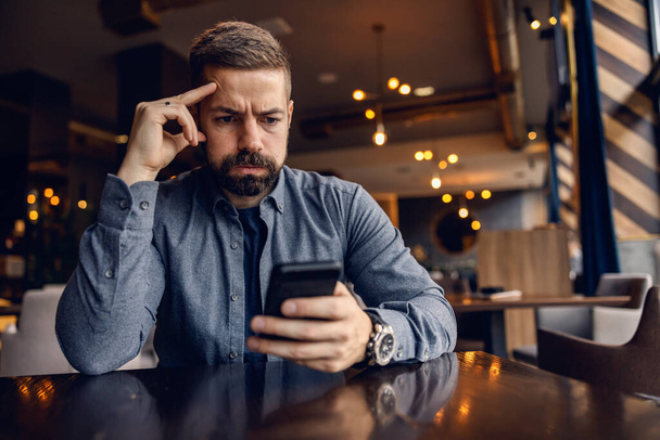 Endişeli bir adam kafeteryada oturuyor ve telefonuna bakıyor.. - Fotoğraf, Görsel