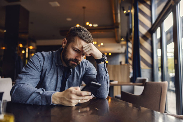 Un joven triste está sentado en la cafetería y mirando su teléfono con desesperación en los ojos. - Foto, Imagen