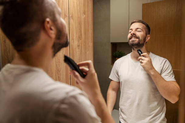 Een gelukkig man houdt de trimmer vast en maakt zich klaar om zijn baard in de badkamer te scheren.. - Foto, afbeelding