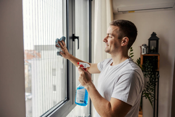 Un uomo ordinato pulisce la finestra con stoffa e detergente a casa. - Foto, immagini