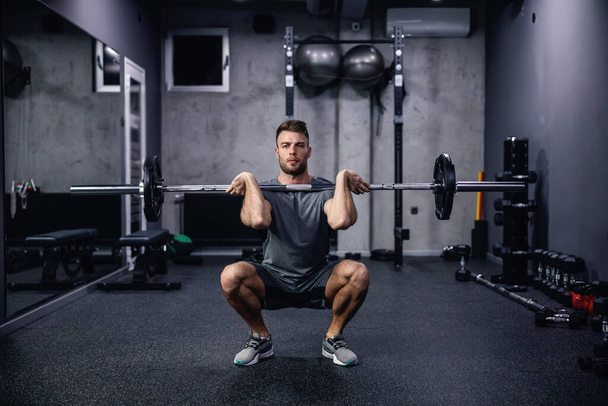 Sports routine and active lifestyle. Front view of a young man in gray sportswear squatting with barbell in an indoor gym with equipment. Fitness body fell good and sexy, an attractive sports guy - Photo, Image